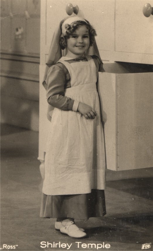 Weiße Kinderschauspielerin (Shirley Temple), die als Krankenschwester gekleidet ist und den Betrachter ansieht.